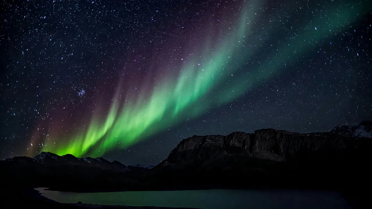 A green and burgundy Northern Lights shines about a dark sky filled with stars over a set of dark shadowed mountains.