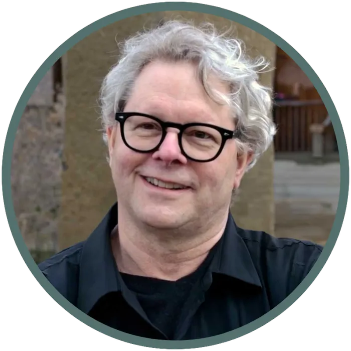 image of a scholarly looking man with wavy gray hair wearing black glasses and a black collared shirt, smiling
